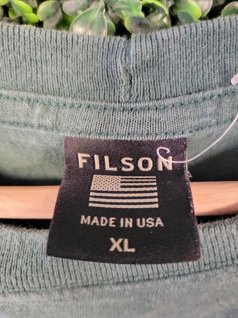 May include: Close-up of a green t-shirt with a black tag. The tag reads "FILSON" above an American flag graphic and "MADE IN USA" with "XL" below.