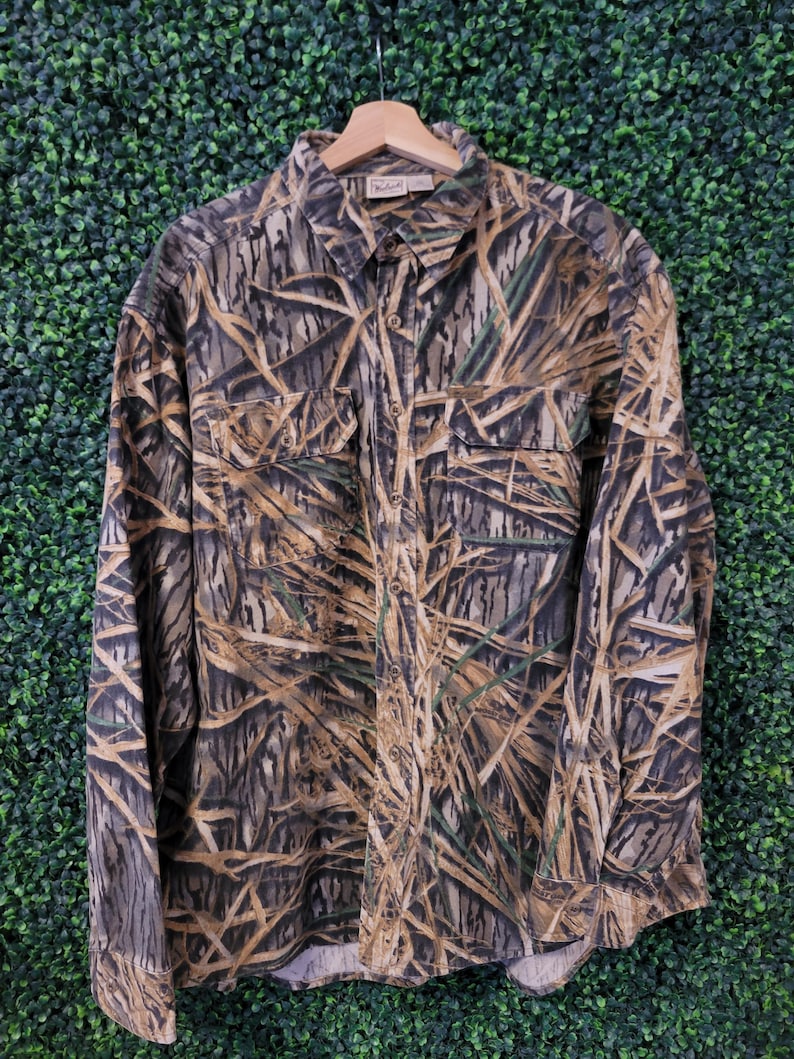 May include: A long-sleeved button-down shirt with a camouflage pattern in shades of brown, green, and tan. The shirt has two chest pockets and is hanging on a wooden hanger. The fabric appears to be a medium-weight material.