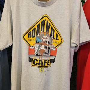 May include: A light gray short-sleeve t-shirt featuring a graphic of a yellow road sign with the text ROADKILL CAFE. The design includes a cartoon deer, a blue bird, and a cat in front of a truck grill with the text YOU KILL IT... WE GRILL IT.