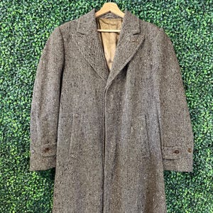 May include: A long, brown and tan herringbone patterned overcoat featuring a notched lapel, a tan interior lining, and two side pockets. The sleeves are finished with buttoned cuffs, and the coat is displayed on a wooden hanger.