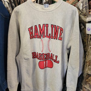 Może przedstawiać: Jasnoszara bluza Hamline Baseball z czerwonymi literami i grafiką baseballową. Numer 11 jest wyświetlany na czerwonym owalu. Bluza wisi na drewnianym wieszaku.