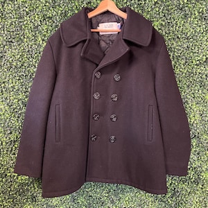 Puede incluir: Un chaquetón cruzado de color marrón oscuro con cuello ancho, dos bolsillos verticales y ocho botones redondos grabados con un diseño de ancla. El forro interior es acolchado y una etiqueta dice U.S. 740N PEA JACKET.