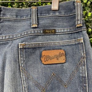May include: Close-up of blue denim jeans with a brown leather patch that reads "Wrangler" on the back pocket. The jeans have a classic five-pocket design with a metal zipper and button closure. The jeans are hanging on a wooden hanger.