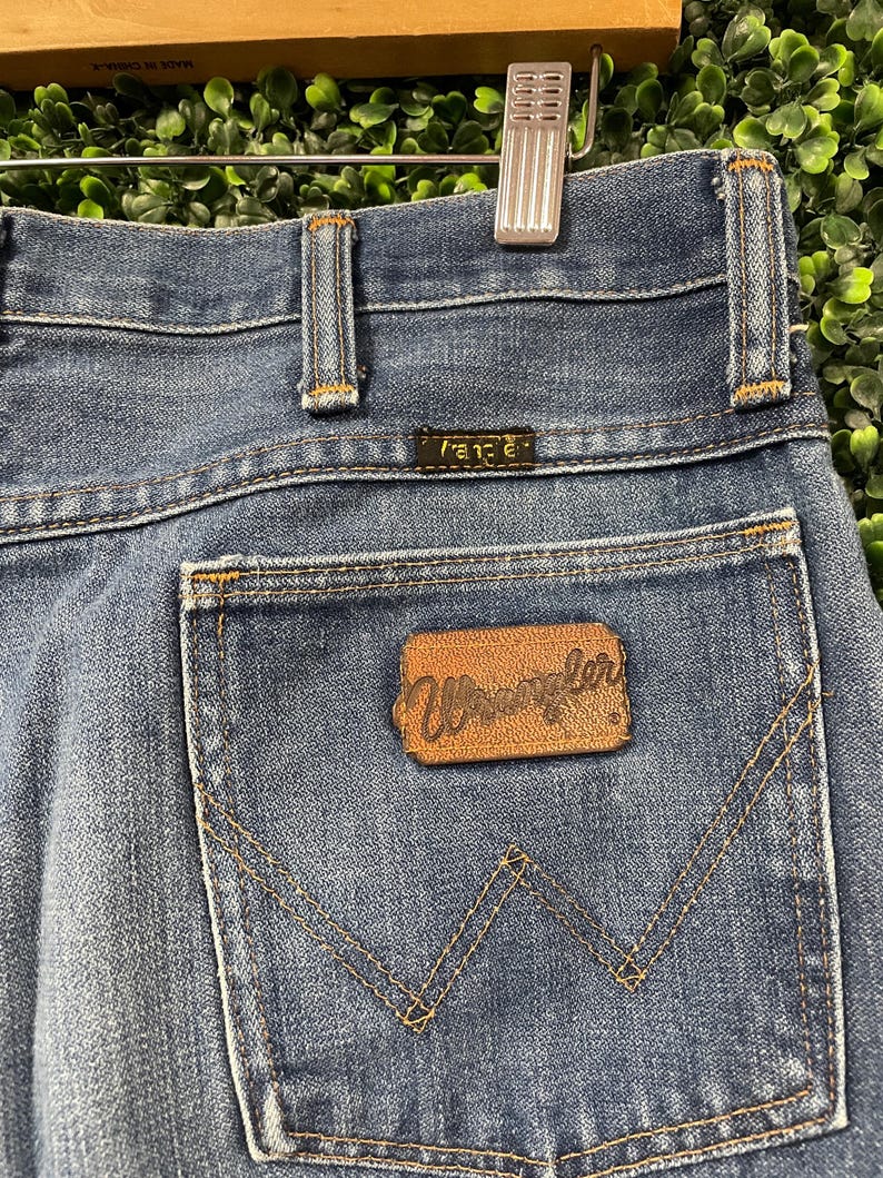 May include: Close-up of blue denim jeans with a brown leather patch that reads "Wrangler" on the back pocket. The jeans have a classic five-pocket design with a metal zipper and button closure. The jeans are hanging on a wooden hanger.