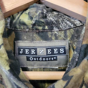 Op de afbeelding: Close-up van een camouflage shirt in groen, bruin en beige, hangend aan een houten hanger. Het label leest "JERZEES Outdoors" in witte tekst op een donkere achtergrond. Geschikt voor buitenactiviteiten.