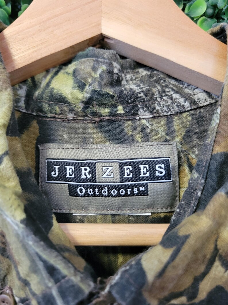 Op de afbeelding: Close-up van een camouflage shirt in groen, bruin en beige, hangend aan een houten hanger. Het label leest "JERZEES Outdoors" in witte tekst op een donkere achtergrond. Geschikt voor buitenactiviteiten.