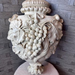 Puede incluir: Jarrón de pared ornamentado de color blanquecino con un diseño detallado de uvas y hojas. La pieza decorativa presenta un cuerpo redondeado, una tapa abovedada y una superficie texturizada. Las uvas y las hojas están esculpidas en alto relieve.