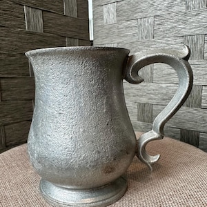 May include: A pewter tankard with a textured surface and a decorative handle. The mug has a flared top and sits on a round base. The handle has a curved design.