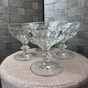 Puede incluir: Cuatro copas de postre transparentes con tallo y un patrón decorativo en el cuenco. Las copas tienen una base redonda y un tallo detallado. Las copas están dispuestas sobre una superficie redonda de color neutro.