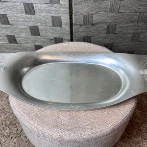May include: A silver-coloured, oval serving tray with handles. The tray has a decorative design on each handle. The tray is sitting on a beige, rounded stool.