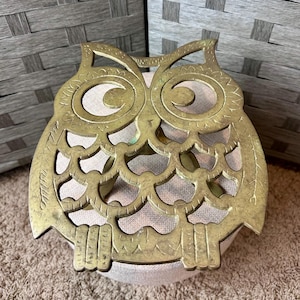 May include: A brass owl-shaped trivet with intricate cut-out details. The trivet has a golden hue and is designed to protect surfaces from hot dishes. The owl's eyes are large and circular, and the wings are spread out.