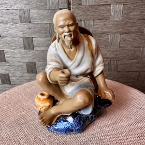 Op de afbeelding: Een keramieken beeldje van een oudere Aziatische man zittend op een blauw-witte basis. Hij draagt een wit gewaad met blauwe accenten en heeft een lange witte baard. Een kleine oranje pot staat naast hem. Het beeldje is gedetailleerd.
