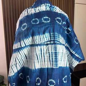 May include: A blue and white tie-dye shirt. The shirt has a button-down collar and short sleeves. The fabric is patterned with white stripes and circular designs against a dark blue background. The shirt is displayed on a mannequin.