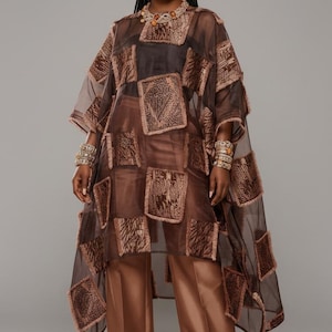 Puede incluir: Una túnica marrón transparente con detalles de patchwork cuadrados, combinada con pantalones de pernera ancha a juego. El conjunto se complementa con joyas doradas, incluyendo un collar, pendientes y pulseras. La modelo lleva zapatos dorados.