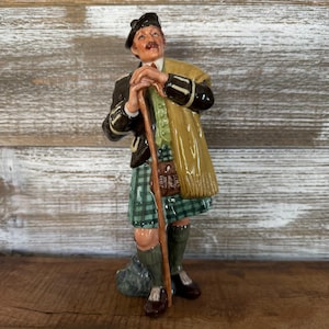 Könnte beinhalten: Eine Keramikfigur einer Person in dunkler Jacke, einem grün-schwarz karierten Kilt und einem beigen Schal. Die Figur trägt eine Schottenmütze, hält einen hölzernen Gehstock und hat eine kleine braune Tasche an der Vorderseite des Kilts befestigt.
