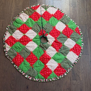 Könnte beinhalten: Ein gesteppter Weihnachtsbaumrock mit Patchwork-Design. Der Rock hat rote, grüne und weiße Quadrate, die jeweils mit einer kleinen roten Schleife gebunden sind. Der Rand ist mit einem Rüschenbesatz versehen, der eine festliche Weihnachtsdekoration schafft.
