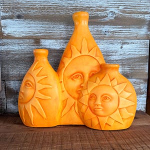 May include: A triple-vessel orange ceramic vase featuring three distinct sun faces with radiating rays. The central vase is the tallest, flanked by two shorter vases on either side. Each sun face has detailed eyes, a nose, and a mouth in relief.