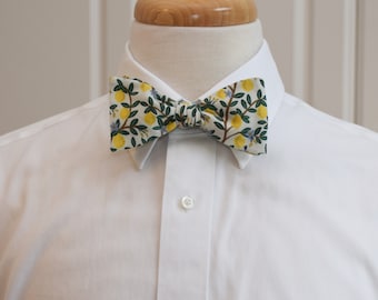 May include: A bow tie with a white background featuring a pattern of yellow lemons, green leaves, and small blue flowers. The bow tie is knotted and displayed on a white dress shirt.
