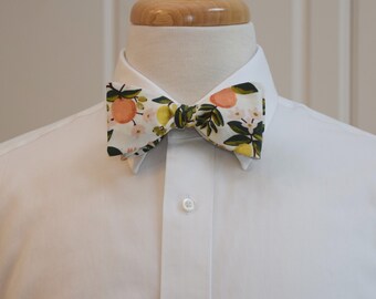 May include: A white bow tie with a colorful floral pattern featuring peaches, lemons, and green leaves. The bow tie is set against a white dress shirt, showcasing the vibrant design. The bow tie is a stylish accessory.