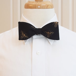 May include: A black bow tie with a repeating gold bee pattern. The bow tie is knotted and worn over a white collared shirt. The bow tie is a formal accessory.