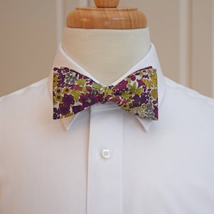 May include: A floral patterned bow tie in shades of purple, yellow, and green is centered on a white dress shirt. The bow tie is the focal point, showcasing a detailed botanical design. The shirt is buttoned at the collar and chest.