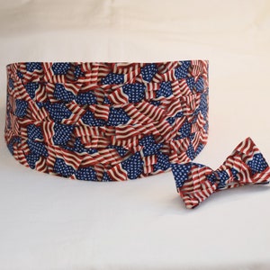 May include: A cummerbund and bow tie set. Both items feature a repeating pattern of the American flag in red, white, and blue. The cummerbund is a wide band, and the bow tie is pre-tied.