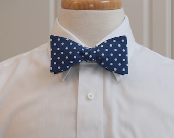 May include: A navy blue bow tie with white polka dots is displayed on a white dress shirt. The bow tie is knotted in the centre and has a classic shape. The shirt has a button-down collar.