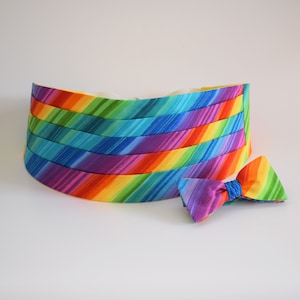 May include: A rainbow-striped cummerbund and matching bow tie set. The cummerbund is a wide band of fabric with diagonal stripes in a spectrum of colors. The bow tie is a small, pre-tied bow with the same rainbow pattern. The set is likely for formal wear.