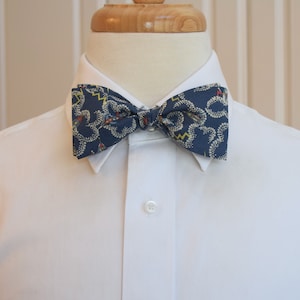 May include: A navy blue bow tie with a repeating pattern of white, yellow, and red shapes. The bow tie is on a white dress shirt, displayed on a mannequin.