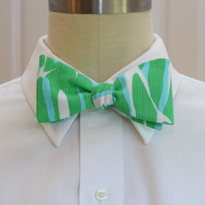 May include: A vibrant green bow tie with a white and light blue abstract pattern. The bow tie is knotted and displayed on a white collared shirt. The design features vertical and curved lines.