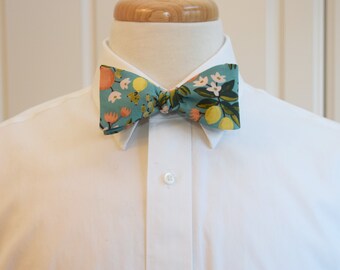 May include: A teal bow tie with a floral pattern featuring lemons, flowers, and oranges. The bow tie is worn with a white dress shirt. The bow tie is a stylish accessory for formal or semi-formal occasions.