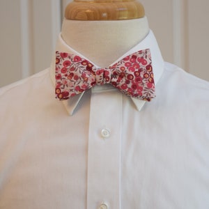 May include: A floral bow tie with a pattern of red and pink flowers on a white background. The bow tie is knotted and positioned on a white collared shirt.