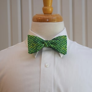 May include: A green bow tie with a scale pattern is displayed on a white collared shirt. The bow tie features shades of green and yellow, creating a textured appearance. The shirt is buttoned at the collar.