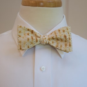 May include: A beige bow tie with a pattern resembling matzah, worn over a white dress shirt. The bow tie has a repeating pattern of small brown dots and a knotted center, creating a unique and festive accessory.