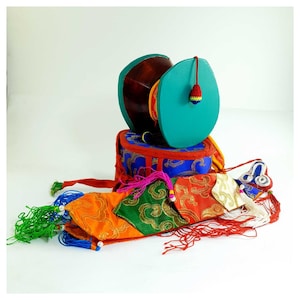 Puede incluir: Un tambor tibetano tradicional con una cara verde turquesa y un cuerpo de madera marrón. El tambor se encuentra sobre una base cilíndrica decorada con detalles rojos, azules y dorados. Bolsas de tela y borlas coloridas están dispuestas alrededor del tambor.
