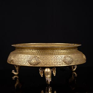 May include: A gold-colored, ornate bowl with a wide, shallow design. The bowl features intricate floral patterns and is supported by three elephant-shaped feet. The rim has a decorative edge. The bowl is likely made of metal.