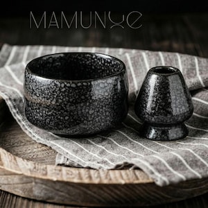 May include: A black ceramic matcha bowl and whisk set with a speckled pattern, on a striped cloth and a wooden tray. The bowl is round, and the whisk has a conical shape. The word "MAMUNYE" is displayed at the top.
