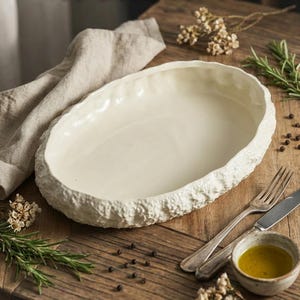 May include: Cream-colored, oval-shaped ceramic serving dish with a textured, stone-like rim. The dish sits on a wooden surface, alongside a linen cloth, herbs, and silverware. The word "MAMUNYE" is displayed at the top of the image.