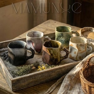 Pode incluir: Uma bandeja de madeira com seis canecas de cerâmica artesanais com texturas e cores únicas, incluindo preto, roxo, verde e castanho. As canecas têm alças e estão acompanhadas de ervas e flores secas, criando uma estética rústica.