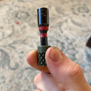 Puede incluir: Un adaptador de enchufe negro y rojo con una base verde texturizada. El adaptador se sostiene en una mano, mostrando su tamaño compacto y diseño. La herramienta parece nueva y sin usar.