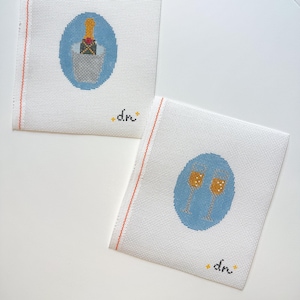 May include: Two white canvas coasters with embroidered designs. One features a champagne bottle in an ice bucket, the other two champagne flutes. Both have a blue oval background and the initials "dn".