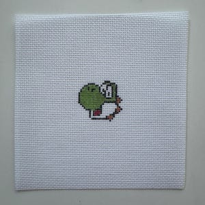Puede incluir: Un lienzo cuadrado de punto de cruz blanco con un personaje Yoshi pixelado. El diseño de Yoshi es principalmente verde con acentos blancos, rojos y marrones. El lienzo tiene una apariencia texturizada y tejida, y la imagen de Yoshi está centrada.