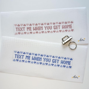 May include: Two white cross-stitch canvases with the text "TEXT ME WHEN YOU GET HOME" in pink and blue, framed by heart patterns. A silver metal clasp and ring are next to the canvases. The canvases have a small signature in the corner.