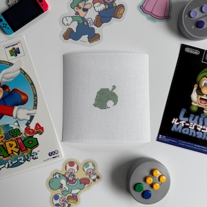 May include: A white square canvas with a green leaf design, surrounded by Nintendo-themed stickers, a game console, and a game cartridge. The canvas features a cross-stitch design of a leaf. The items are arranged on a white surface.