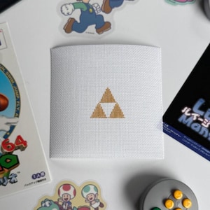 Puede incluir: Cuadrado blanco de punto de cruz con un diseño Trifuerza dorado. El diseño está centrado en la tela. La imagen está rodeada de pegatinas temáticas de videojuegos y un cartucho de juego.