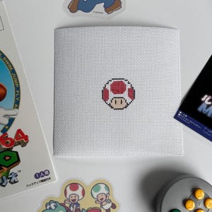 May include: A white square cross-stitch featuring a red and white mushroom design. The cross-stitch is surrounded by video game-themed stickers and a game controller, suggesting a gaming or retro theme.