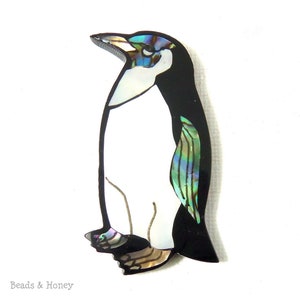 Peut inclure: Broche en forme de pingouin, corps noir, poitrine blanche et détails irisés. La broche présente un effet nacré sur la tête, l'aile et les pieds. Le texte "Beads & Honey" est en bas à gauche.