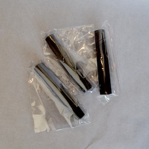 May include: Three black, cylindrical objects, possibly pen parts or similar components, are individually packaged in clear plastic. The items are arranged on a light-colored surface, suggesting a product display or storage setting.
