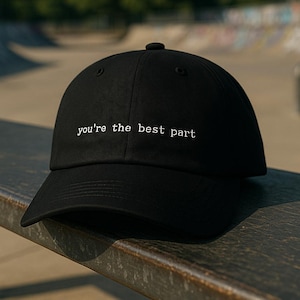 Puede incluir: Gorra de béisbol negra con la inscripción bordada en blanco "you're the best part". La gorra está colocada sobre una superficie oscura y desgastada, con un parque de skate borroso en el fondo.