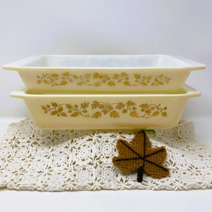 May include: Two rectangular, vintage Pyrex baking dishes. The dishes are a pale yellow with a gold leaf and acorn pattern around the rim. They are stacked on top of each other. A crocheted doily and a brown leaf decoration are in the foreground.